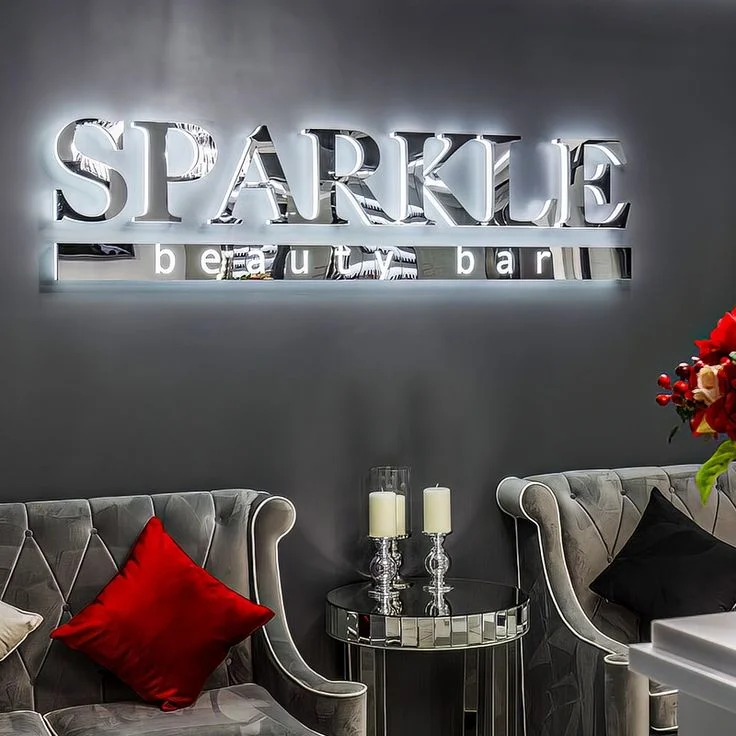 Backlit sign "SPARKLE beauty bar" in mirrored letters on a dark gray wall with elegant decor including plush chairs, a mirrored table, and candles.