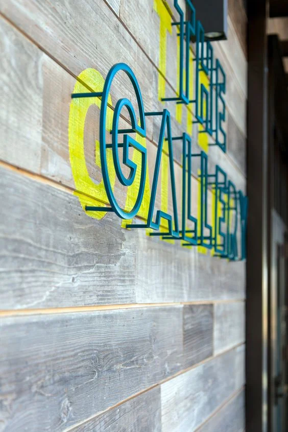 Three-dimensional blue and yellow sign reading "THE GALLERY" mounted on a rustic wooden plank wall.