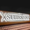 Backlit sign "THE SUITES SPOT" with a scissors icon, mounted on a textured wall above a roof with terracotta tiles, illuminated against a dark night sky.