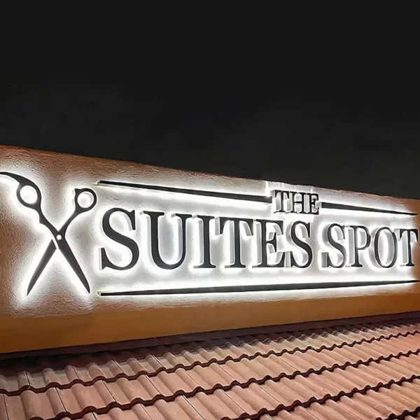 Backlit sign "THE SUITES SPOT" with a scissors icon, mounted on a textured wall above a roof with terracotta tiles, illuminated against a dark night sky.