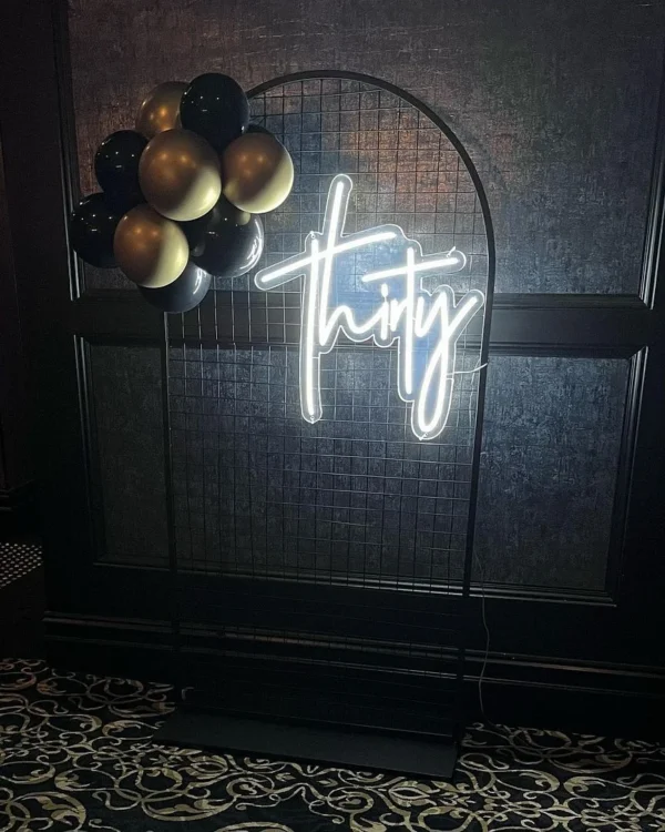 Black and gold balloon arrangement with a white neon sign reading "thirty" on a wireframe backdrop.