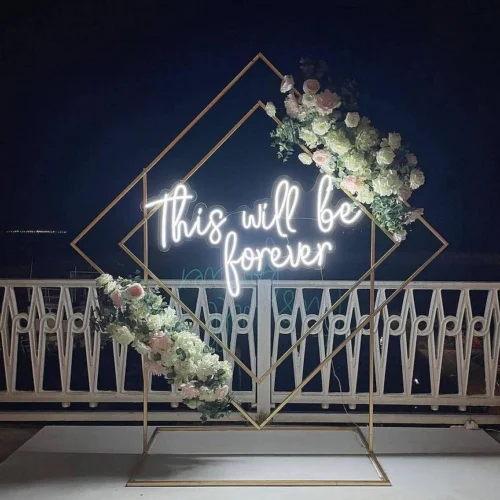 White neon sign reading "This will be forever" in cursive font, mounted on a geometric frame with white and pink flowers, placed on a balcony with a dark night sky backdrop.