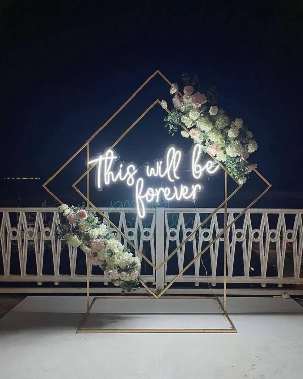 White neon sign reading "This will be forever" in cursive font, mounted on a geometric frame with white and pink flowers, placed on a balcony with a dark night sky backdrop.