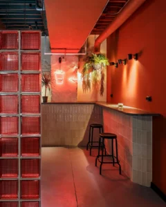 Urban café interior with a red neon coffee cup sign, bar counter with stools, glass block wall, and hanging plants.