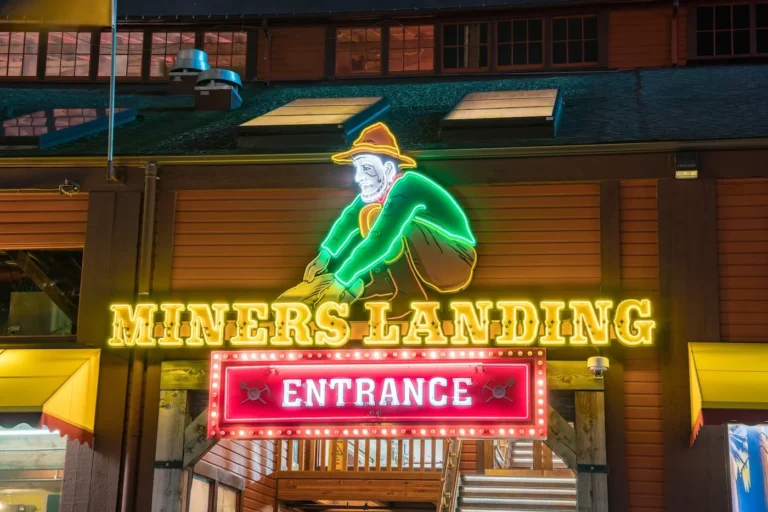 Neon sign for "Miners Landing" featuring a colorful depiction of a miner in a hat crouching and panning for gold. Below the miner, the words "MINERS LANDING" are illuminated in bright yellow neon, and a red neon sign with the word "ENTRANCE" is outlined with bulbs. The sign is mounted on a wooden building facade.