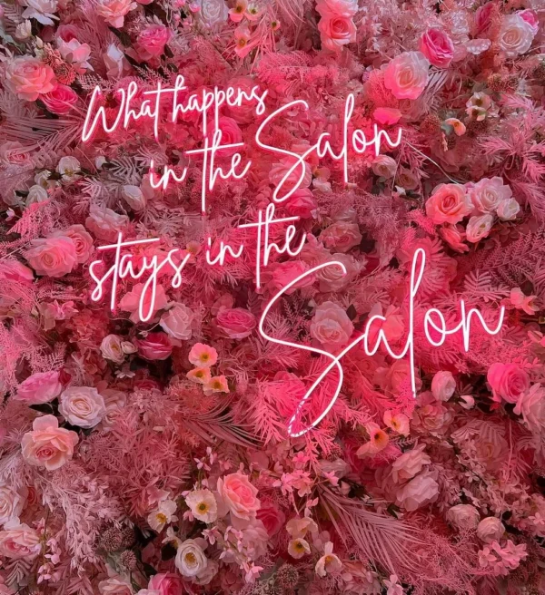 Neon sign stating "What happens in the Salon stays in the Salon" in pink cursive letters, mounted on a wall covered with pink flowers.