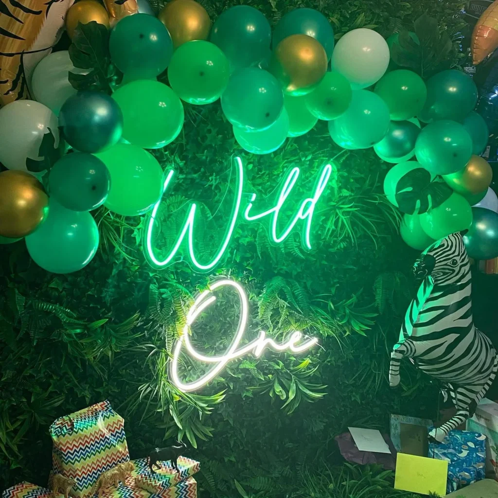 Neon sign with "Wild One" written in bright green and white, surrounded by green and gold balloons, mounted on a lush greenery wall with a zebra decoration.